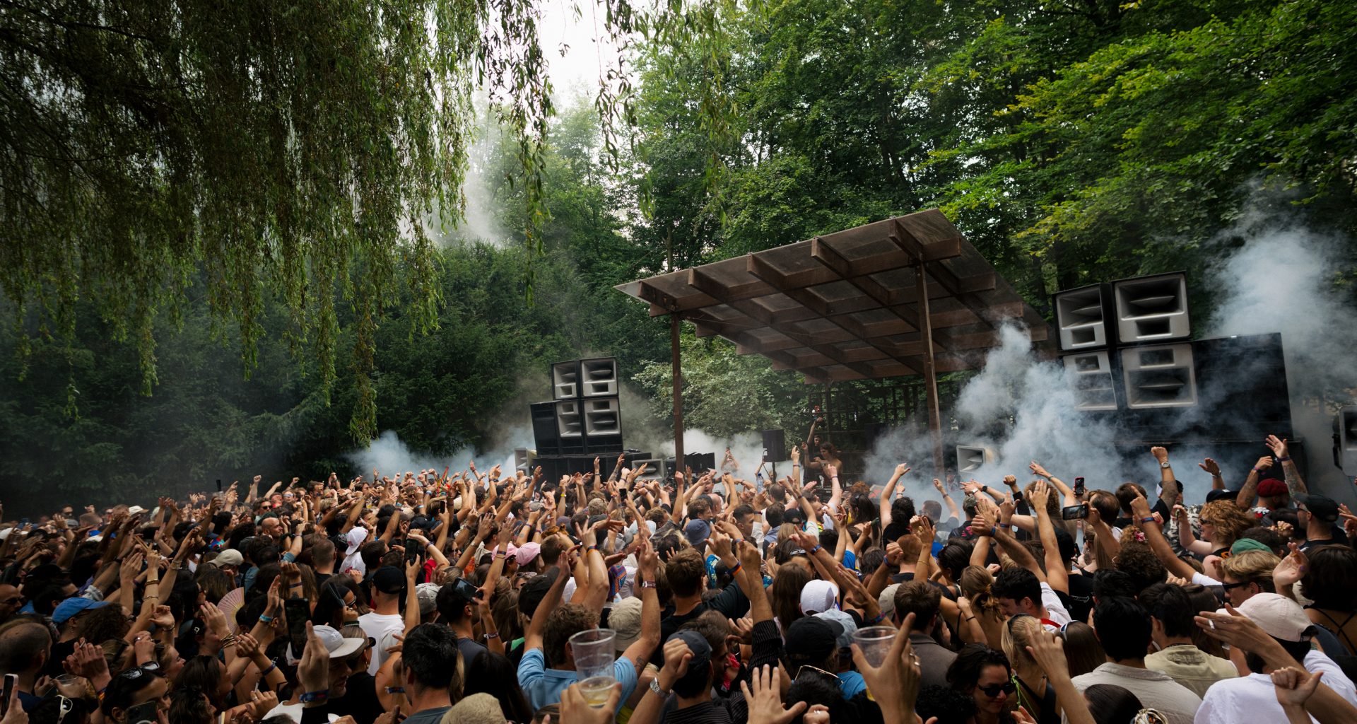 A large crowd dances energetically under the canopy of trees at Dekmantel 2024, creating an immersive festival atmosphere