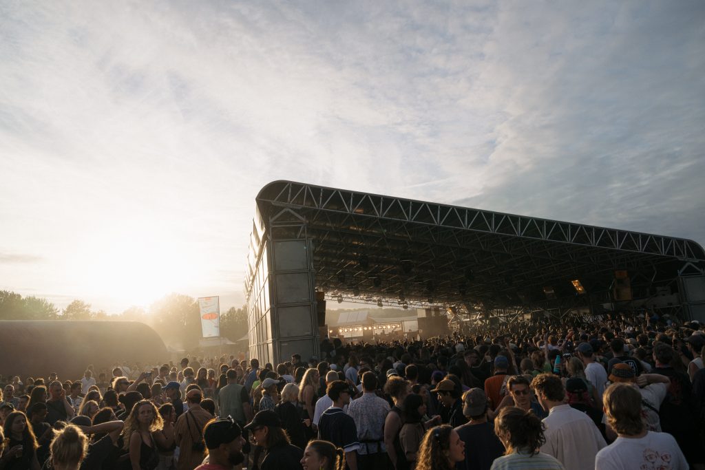 The sun sets over a massive outdoor stage at Dekmantel 2024, where a crowd of festival-goers enjoy the music.