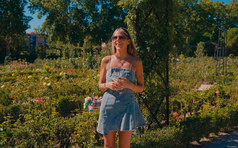 Woman standing outdoors in a garden, smiling during an interview Otta, wearing a denim dress and sunglasses