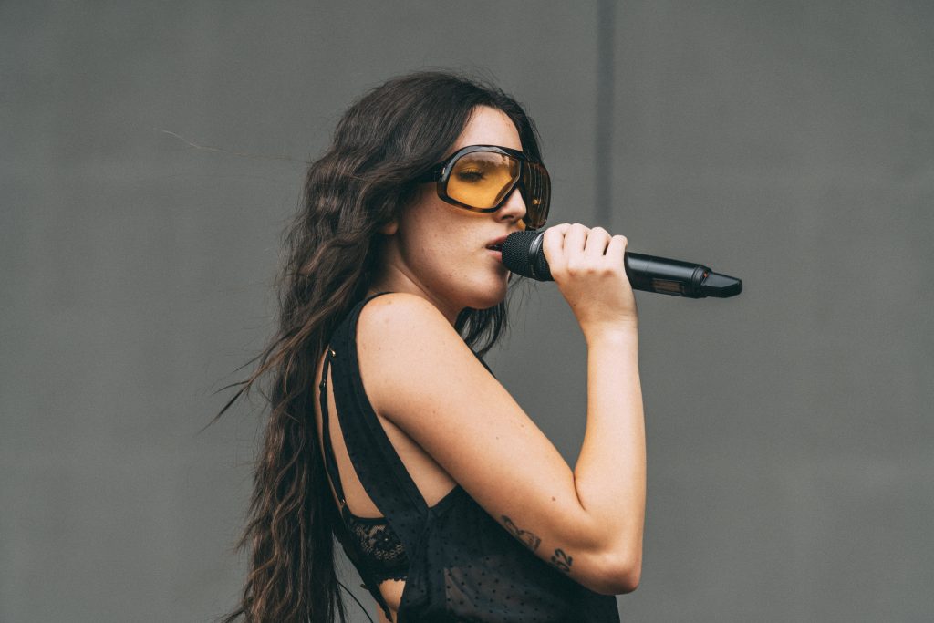 Judeline singing into a microphone during her live performance at Kalorama Madrid, wearing large yellow-tinted sunglasses and a black outfit, captured in a close-up shot