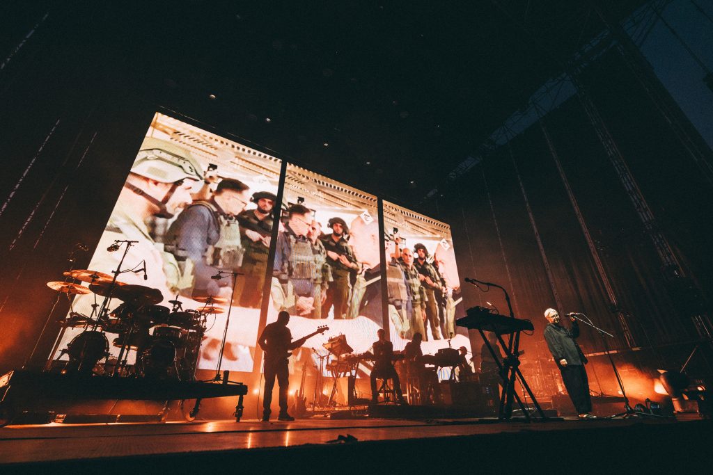 Massive Attack performing live at Kalorama 2024 with a visually striking backdrop featuring military imagery, powerful lighting effects, and a moody atmosphere