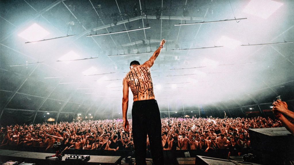 OGUZ standing on stage with his back to the camera, raising his fist in front of a massive crowd at a packed indoor venue