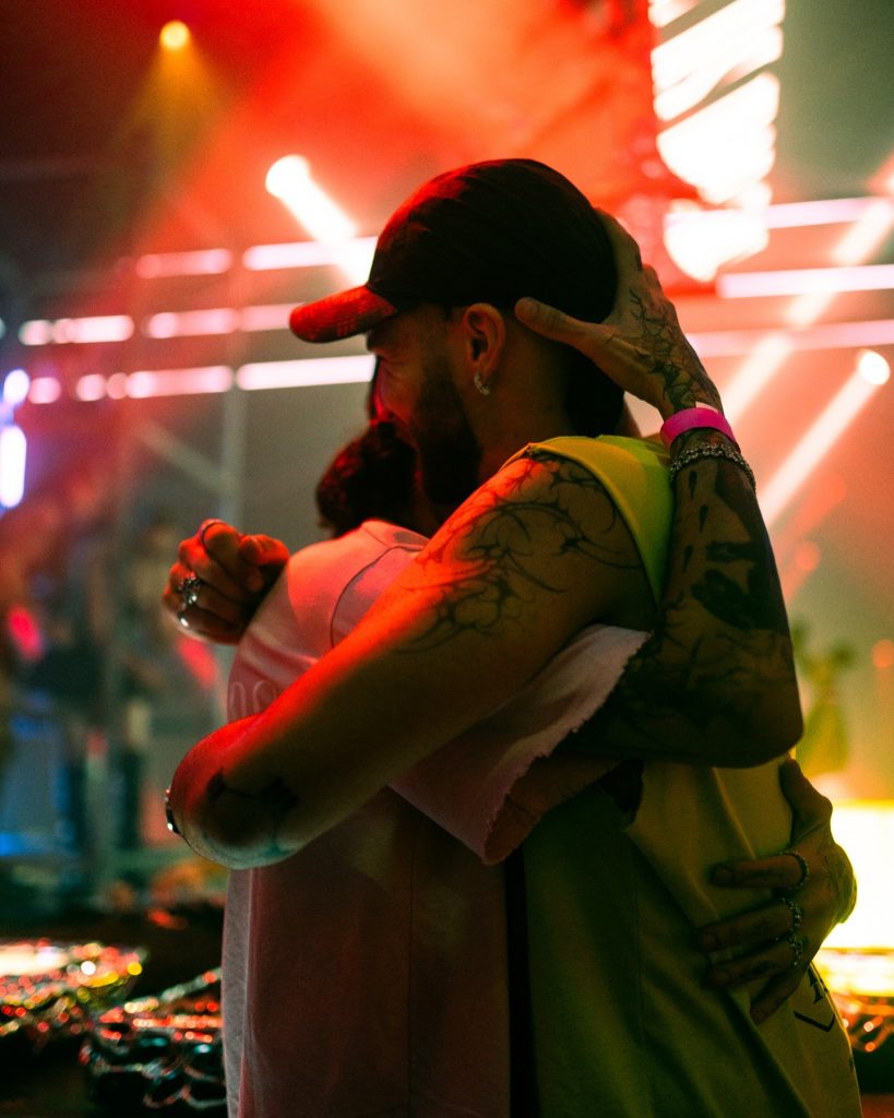 interview OGUZ sharing an emotional embrace with a fellow artist backstage, surrounded by vibrant red and yellow stage lights