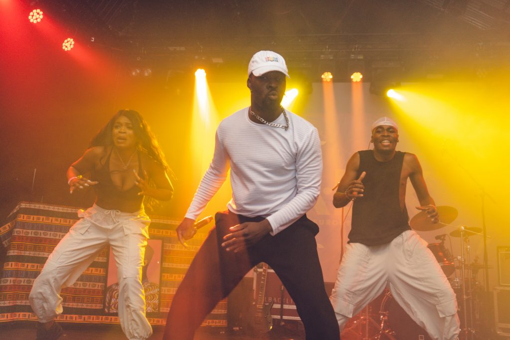 Three dancers perform energetically on stage, illuminated by bright yellow and red stage lights. The lead dancer in a white cap and shirt takes center stage, while the others dance in rhythm, creating a vibrant and dynamic scene at AEX Festival