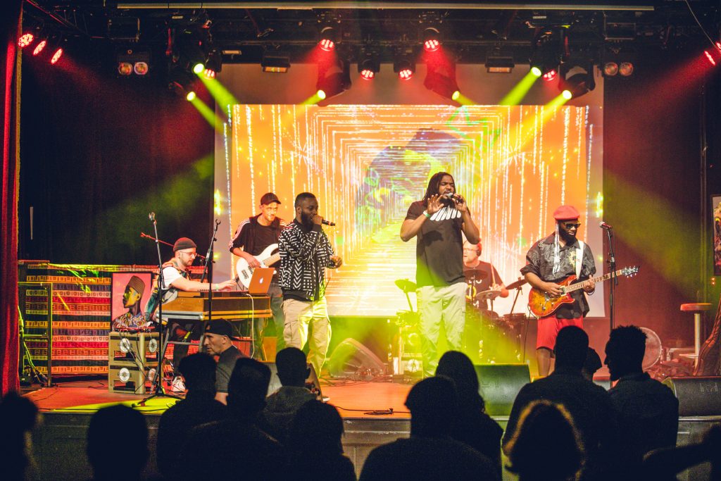 A live band performs on stage with bright, colorful lighting and a digital backdrop. Two vocalists engage with the crowd while the band, including a guitarist, keyboardist, and bassist, plays in the background at AEX Festival