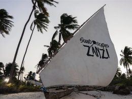 A traditional wooden dhow with a white sail featuring the words 'Soundscapes Zanzi' in bold black letters, set against a backdrop of tall, swaying palm trees on a tropical beach during sunset.