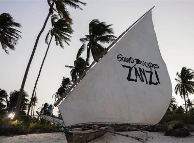 A traditional wooden dhow with a white sail featuring the words 'Soundscapes Zanzi' in bold black letters, set against a backdrop of tall, swaying palm trees on a tropical beach during sunset.