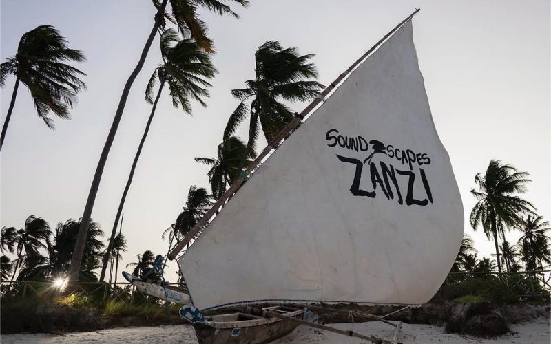 A traditional wooden dhow with a white sail featuring the words 'Soundscapes Zanzi' in bold black letters, set against a backdrop of tall, swaying palm trees on a tropical beach during sunset.