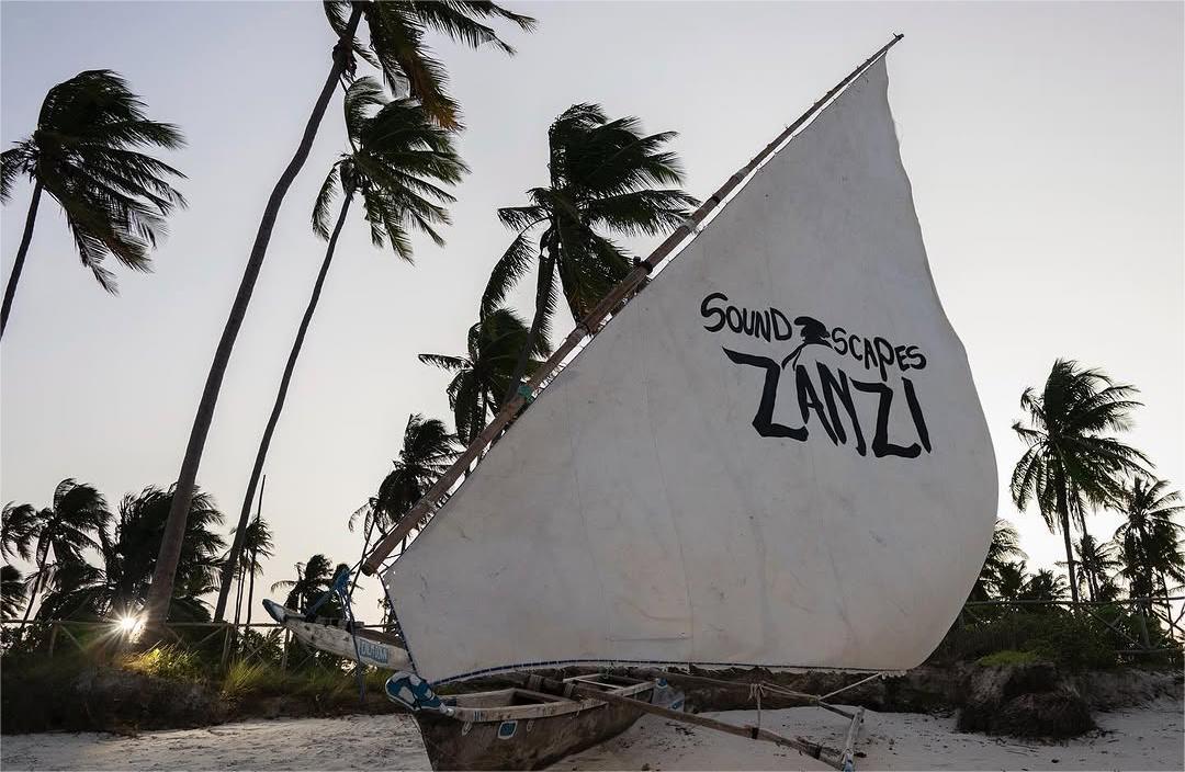 A traditional wooden dhow with a white sail featuring the words 'Soundscapes Zanzi' in bold black letters, set against a backdrop of tall, swaying palm trees on a tropical beach during sunset.